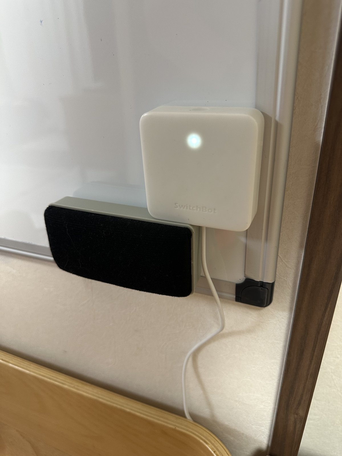 リビングのホワイトボードの隅に取り付けたSwitchBotハブミニ。白い四角い本体のランプが光り、下からケーブルが伸びている
